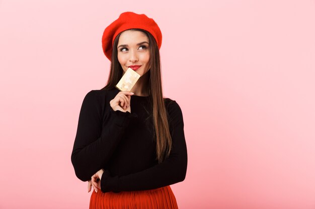 Retrato de uma jovem bonita e feliz, usando uma boina vermelha em pé, isolado sobre um fundo rosa, segurando um cartão de crédito de plástico