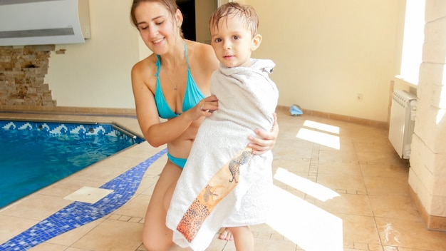 Retrato de uma jovem bonita e sorridente, cobrindo e limpando seu filho de 3 anos de idade após nadar na piscina coberta
