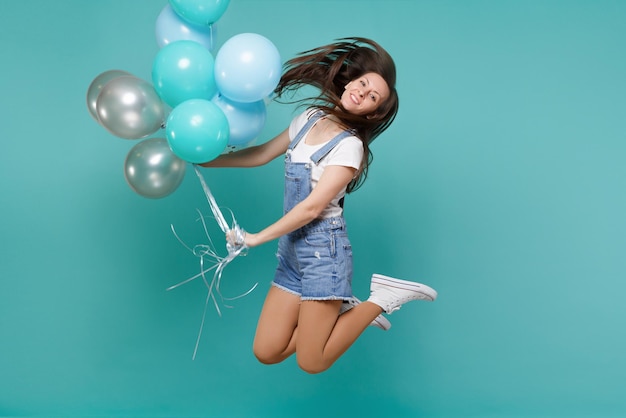 Retrato de uma jovem bonita em roupas jeans pulando alto, comemorando e segurando balões de ar coloridos isolados no fundo da parede azul turquesa. Festa de aniversário, conceito de emoções de pessoas.