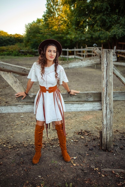 Retrato de uma jovem bonita em um vestido branco com botas acima do joelho e um chapéu com um cinto e uma franja em um estábulo de fazenda rústica