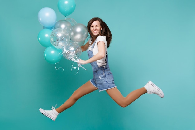 Retrato de uma jovem bonita feliz em roupas jeans pulando alto, comemorando e segurando balões de ar coloridos isolados em fundo azul turquesa. Festa de aniversário, conceito de emoções de pessoas.