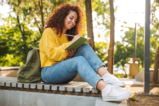 Retrato de uma jovem bonita feliz estudante encaracolado sentado ao ar livre no parque natural, escrevendo notas no caderno.