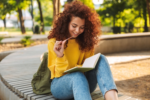 Retrato de uma jovem bonita feliz estudante encaracolado sentado ao ar livre no parque natural, escrevendo notas no caderno.