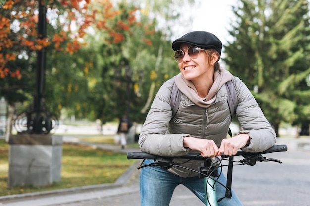 Retrato de uma jovem bonita na moda em boné e óculos de sol em bicicleta no dia ensolarado de outono no parque da cidade