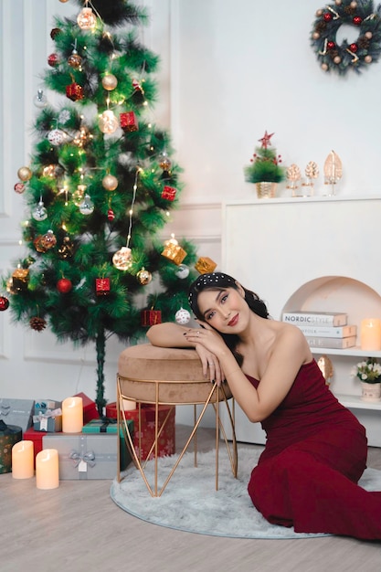 Retrato de uma jovem bonita sentada no chão sorrindo usando um vestido vermelho na sala de estar decorada de Natal dentro de casa