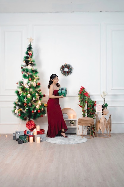 Retrato de uma jovem bonita sentada segurando um presente de Natal sorrindo usando um vestido vermelho na sala de estar decorada de Natal dentro de casa