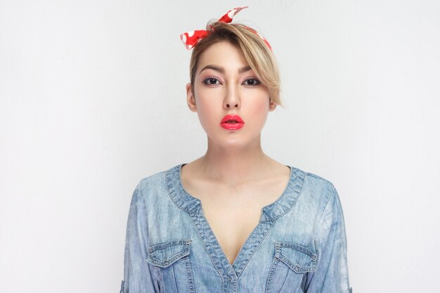 Retrato de uma jovem bonita séria calma em camisa jeans azul casual com maquiagem e lábios vermelhos e bandana em pé e olhando para a câmera. tiro do estúdio interno, isolado no fundo branco.