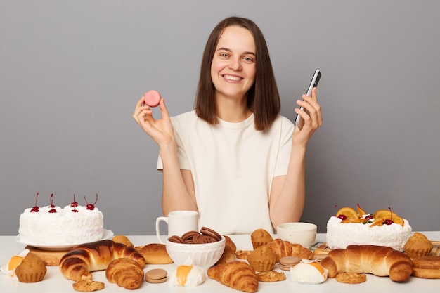 Retrato de uma jovem bonita sorridente com cabelos castanhos sentado à mesa com muita massa segurando biscoitos deliciosos e telefone inteligente pedindo padaria on-line isolada sobre fundo cinza