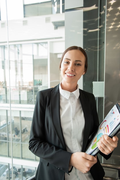 Retrato de uma jovem bonita sorridente com uma jaqueta segurando papéis milimetrados e de pé no elevador do escritório