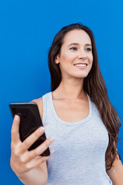 Retrato de uma jovem bonita sorrindo e usando um smartphone com uma parede azul ao fundo