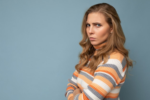 Retrato de uma jovem bonita, triste com raiva insatisfeita loira com emoções sinceras, vestindo uma blusa listrada casual isolada sobre um fundo azul com espaço de cópia