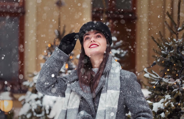 Retrato de uma jovem bonita usa chapéu preto estiloso se perguntando pela neve antes do fundo da cidade de Natal na época da neve