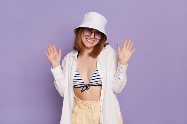 Retrato de uma jovem bonita vestindo camisa branca do panamá e maiô posando isolado sobre fundo roxo em pé com os braços levantados e sorriso cheio de dentes