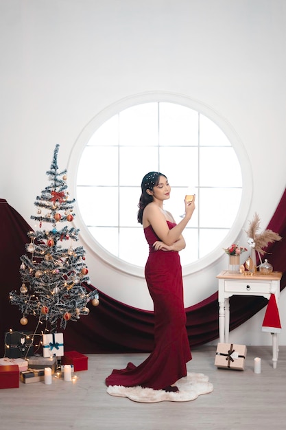 Retrato de uma jovem bonita vestindo um vestido vermelho e bebendo vinho sorrindo para a câmera em pé na sala de estar decorada de Natal dentro de casa