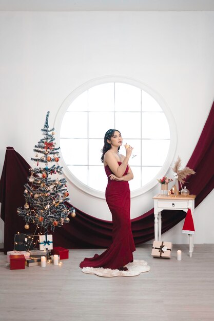 Retrato de uma jovem bonita vestindo um vestido vermelho e bebendo vinho sorrindo para a câmera em pé na sala de estar decorada de Natal dentro de casa