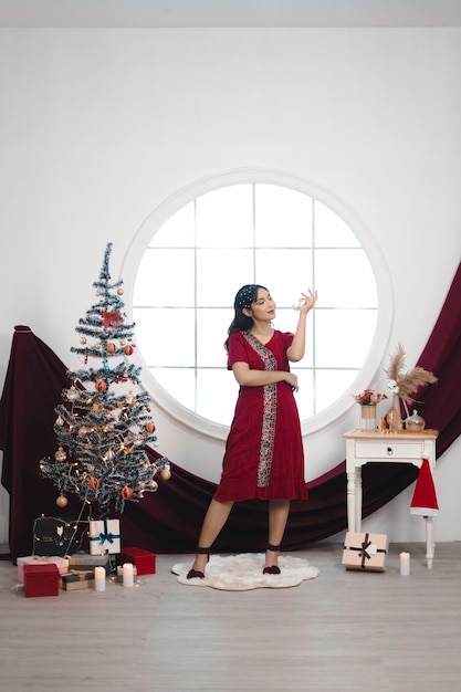 Foto retrato de uma jovem bonita vestindo um vestido vermelho sorrindo para a câmera em pé na sala de estar decorada de natal dentro de casa