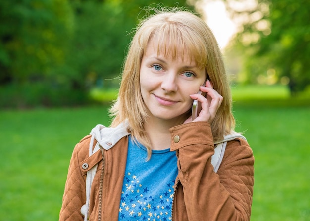 Retrato de uma jovem caucasiana atraente e sorridente com celular em Spring Park olhando para a câmera de perto.