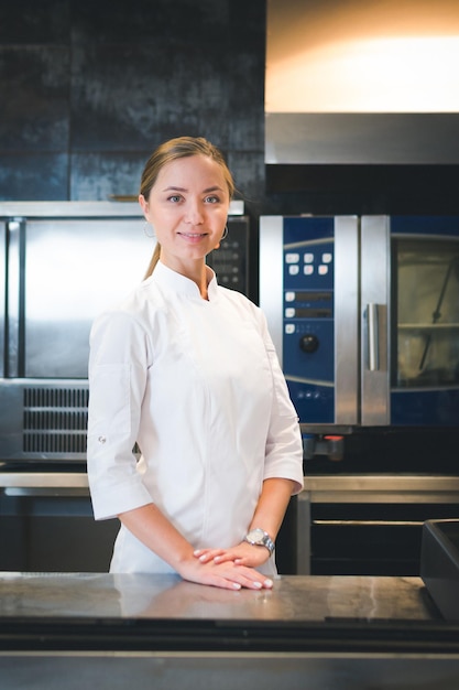 Retrato de uma jovem chef confiante e sorridente, vestida de cozinha profissional uniforme branca, está em segundo plano