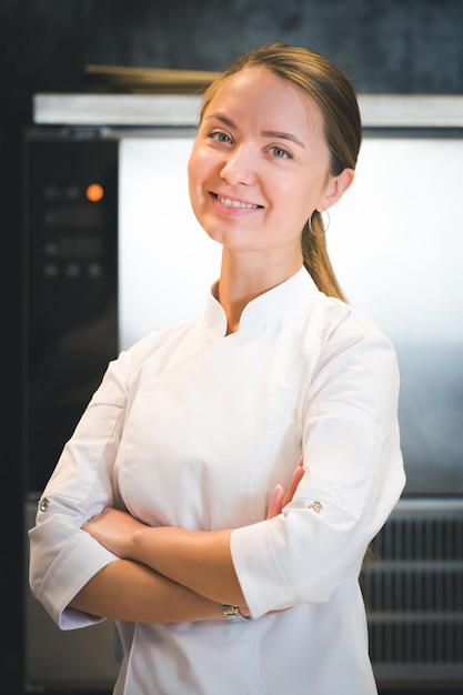 Retrato de uma jovem chef confiante e sorridente, vestida de cozinha profissional uniforme branca, está em segundo plano