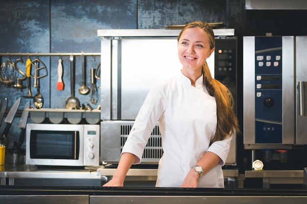 Retrato de uma jovem chef confiante e sorridente, vestida de cozinha profissional uniforme branca, está em segundo plano