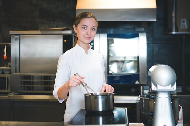 Retrato de uma jovem chef confiante e sorridente, vestida de cozinha profissional uniforme branca, está em segundo plano