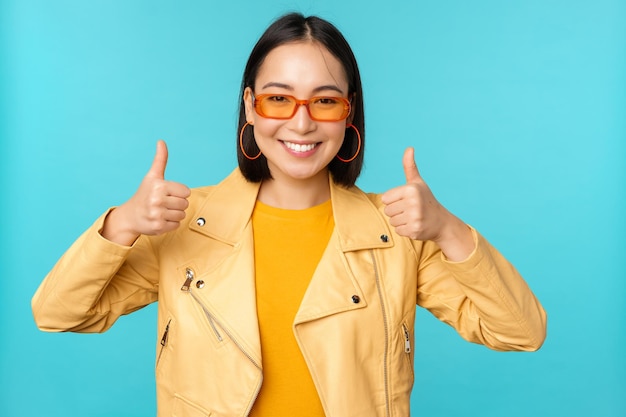 Retrato de uma jovem chinesa asiática moderna em óculos de sol usa roupas da moda mostrando os polegares para cima e sorrindo satisfeito recomendando smth fundo azul
