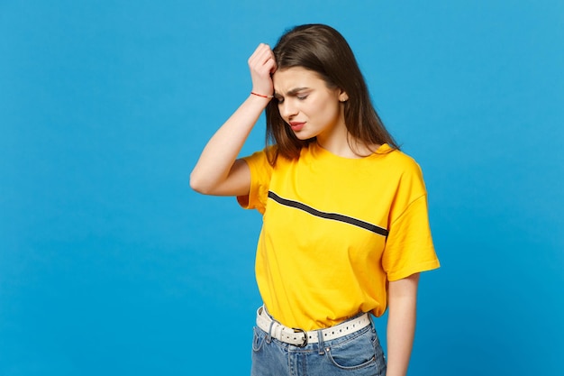 Retrato de uma jovem chorando descontente em roupas casuais vívidas, mantendo os olhos fechados, colocando a mão na cabeça isolada em fundo azul brilhante no estúdio. Conceito de estilo de vida de pessoas. Mock up espaço de cópia.