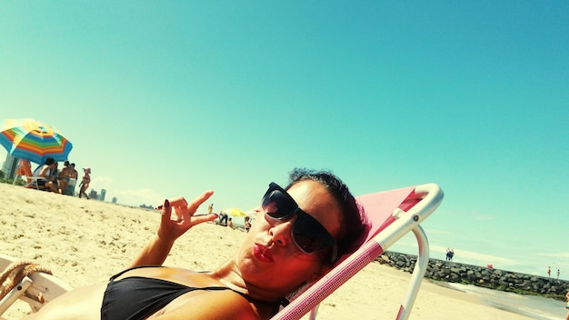 Retrato de uma jovem com óculos de sol relaxando em uma cadeira na praia contra o céu azul