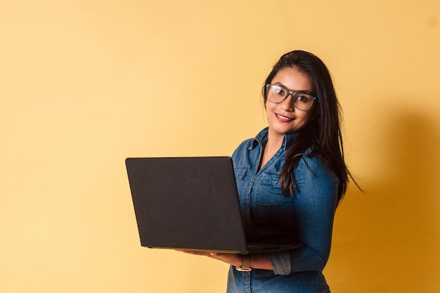 Retrato de uma jovem com óculos segurando laptop sorrindo olha a câmera sobre fundo amarelo