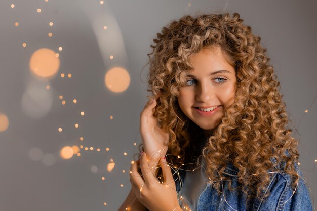 Retrato de uma jovem com olhos azuis e cachos segurando luzes brilhantes de uma guirlanda em suas mãos