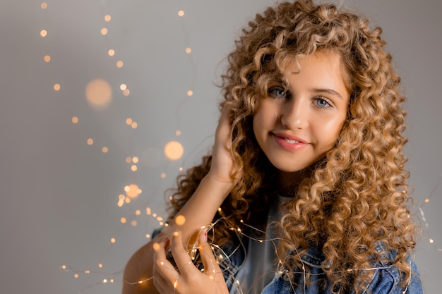 Retrato de uma jovem com olhos azuis e cachos segurando luzes brilhantes de uma guirlanda em suas mãos