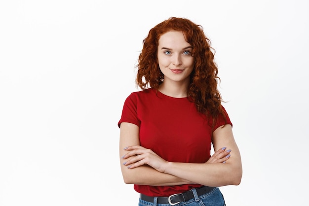 Retrato de uma jovem confiante com cabelo encaracolado ruivo em pé autoconfiante com os braços cruzados no peito sorrindo para a câmera em pé na camiseta contra o fundo branco