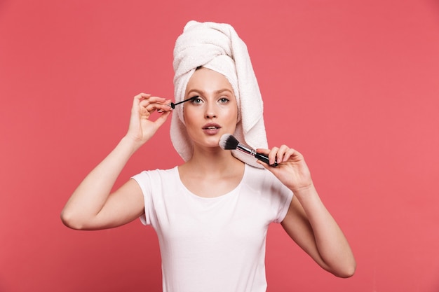 Retrato de uma jovem contente enrolada em uma toalha branca após o banho, aplicando cosméticos com um pincel de maquiagem isolado sobre a parede rosa