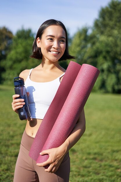 Retrato de uma jovem coreana magra e saudável fazendo exercícios no parque em pé com garrafa de água e