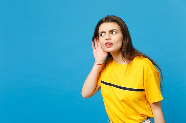 Retrato de uma jovem curiosa em roupas casuais vívidas, olhando de lado, tente ouvi-lo com a mão perto da orelha isolada no fundo da parede azul no estúdio. Conceito de estilo de vida de pessoas. Mock up espaço de cópia.