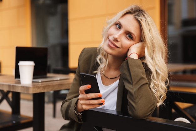 Retrato de uma jovem de 20 anos segurando um smartphone, enquanto está sentado em um café ao ar livre com um café para viagem e um laptop prateado