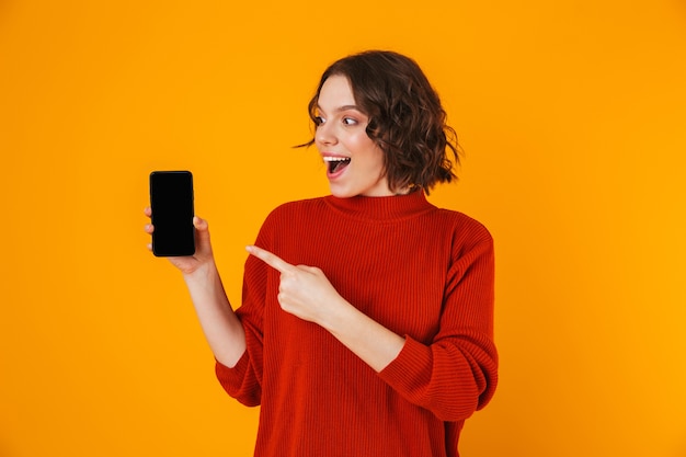 Retrato de uma jovem de 20 anos vestindo um suéter segurando e demonstrando o smartphone em pé isolado sobre o amarelo