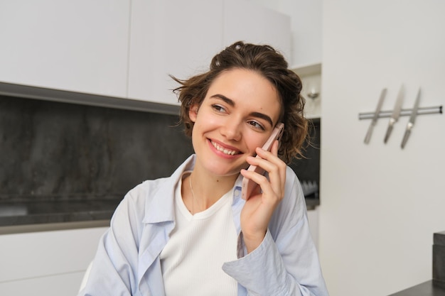 Retrato de uma jovem de anos segurando um smartphone falando no celular sentado na cozinha em