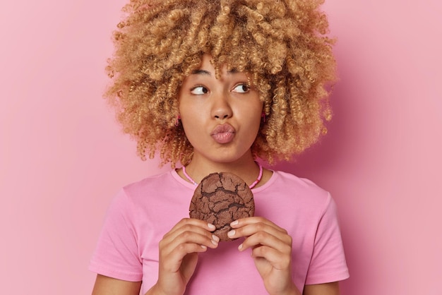 Retrato de uma jovem de cabelos encaracolados bonita mantém os lábios dobrados segura biscoito de chocolate delicioso quer comer comida doce vestida com camiseta casual isolada sobre fundo rosa Hora do lanche