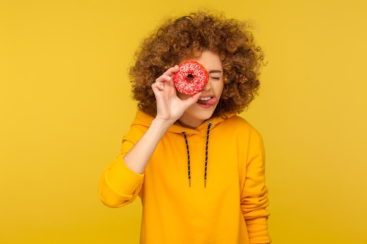 Foto retrato de uma jovem de cabelos encaracolados curiosa positiva com capuz estilo urbano olhando através de rosquinha e mostrando a língua, se divertindo, brincando com rosquinha açucarada. estúdio interno tiro fundo amarelo