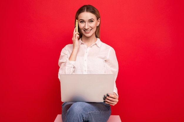 Retrato de uma jovem de camisa branca falando ao telefone e segurando um laptop em um fundo vermelho