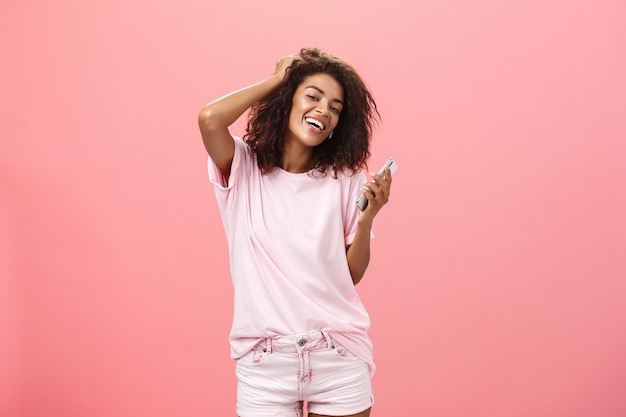 Retrato de uma jovem de pele escura, moderna e despreocupada, usando um smartphone, tocando o cabelo com alegria e olhando com um largo sorriso, segurando o celular posando contra a parede rosa