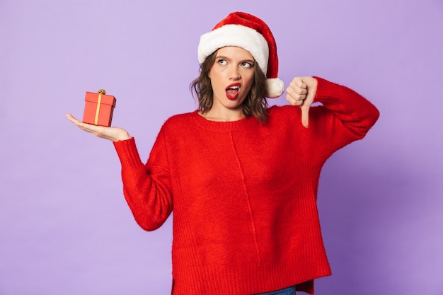 Retrato de uma jovem descontente com chapéu de Natal, isolado na parede roxa, segurando uma caixa de presente surpresa.