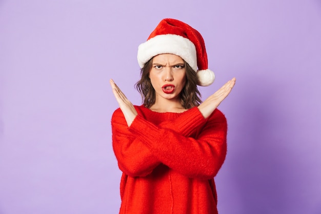 Retrato de uma jovem descontente com chapéu de natal isolado sobre a parede roxa, mostrando o gesto de parada.