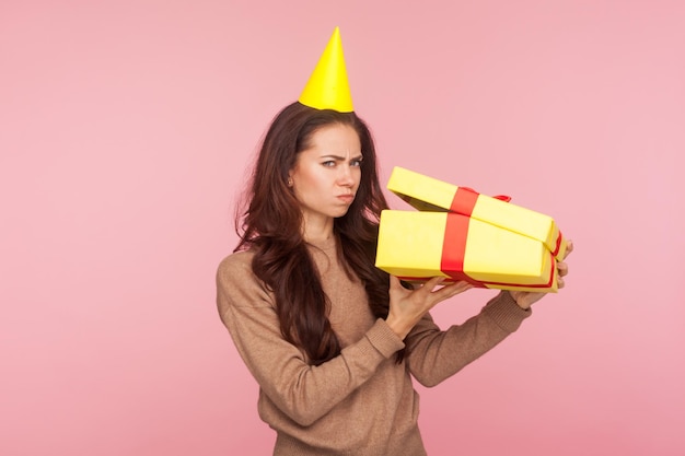 Retrato de uma jovem descontente engraçada com cone de festa na cabeça desembalando o presente e parecendo desapontado chateado com o erro de entrega de presente de aniversário tiro de estúdio interno isolado no fundo rosa