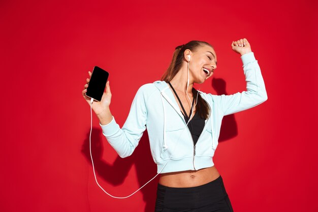 Retrato de uma jovem desportista em forma bonita, de pé, a ouvir música com auscultadores, a segurar num telemóvel