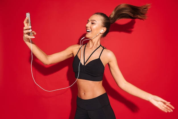 Retrato de uma jovem desportista em forma bonita, pulando, ouvindo música com fones de ouvido, segurando o telefone celular