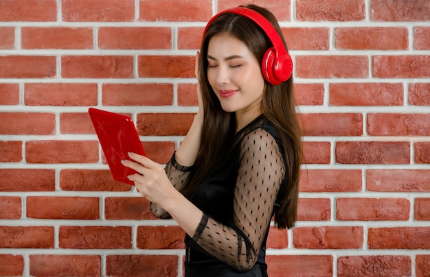 Retrato de uma jovem e atraente modelo feminino asiático em preto revelando vestido usando fone de ouvido vermelho, ouvir música e segurando o tablet contra o fundo da parede de tijolo laranja.