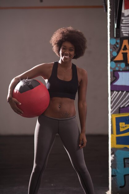 Retrato de uma jovem e bela mulher afro-americana carregando bola crossfit no ginásio