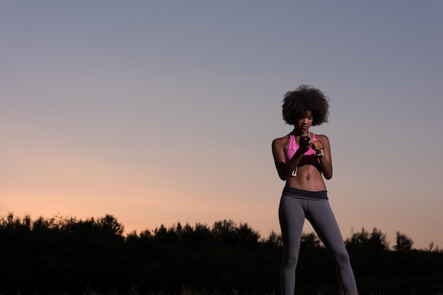 retrato de uma jovem e bela mulher afro-americana com fones de ouvido e celular correndo na natureza linda noite de verão